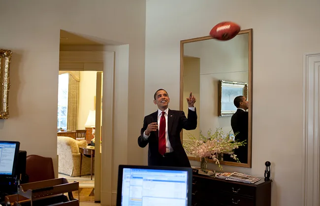 Man in a suit tossing a football inside an office, reflected in a mirror; computers and floral arrangement visible.