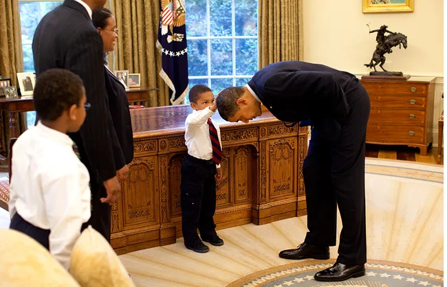A boy touches a man's head in an elegant office with family and ornate desk nearby.