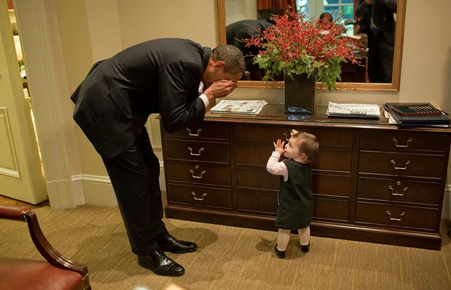 A man bends over to play peekaboo with a toddler in an office setting, with a mirror and flowers in the background.