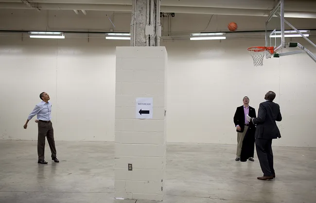 Three men play basketball indoors near a brick column; one shoots, two watch, with a "motorcade" sign on the wall.