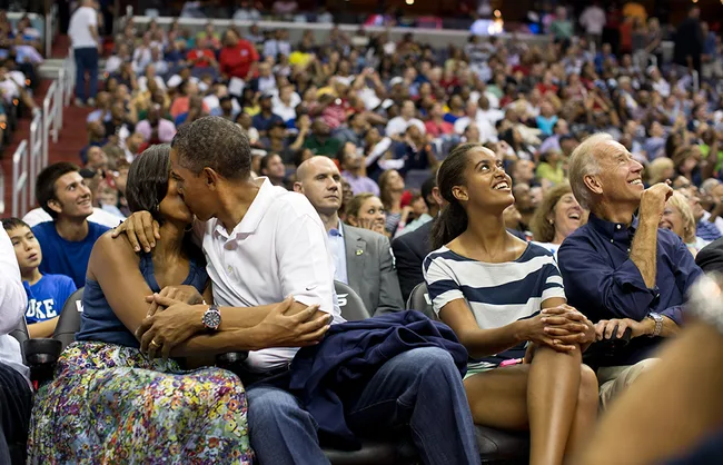 A couple kisses while two others sit and smile at a basketball event, with a cheering crowd in the background.