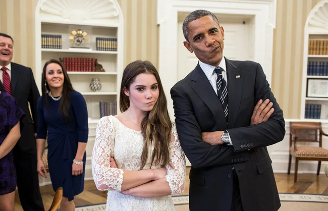 Two individuals stand with arms crossed, mimicking each other's expression, in a formal setting, smiling people behind.