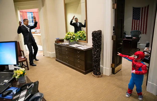 Child in Spider-Man costume playfully points at a smiling man in an office with hands raised in pretend surrender.