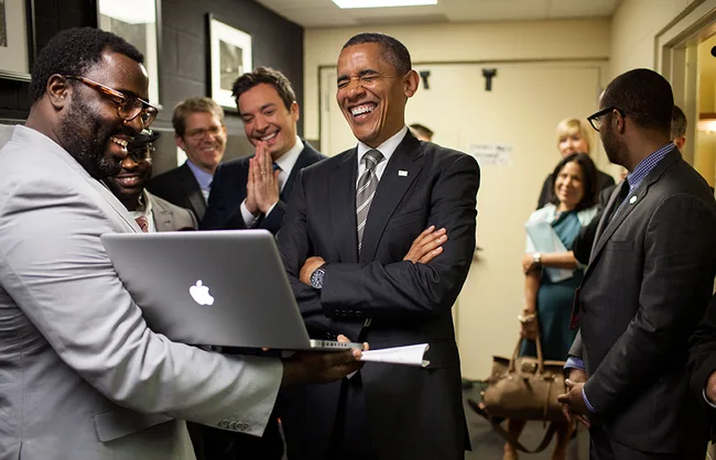 Group of people in suits laughing and smiling while looking at a laptop in a hallway.