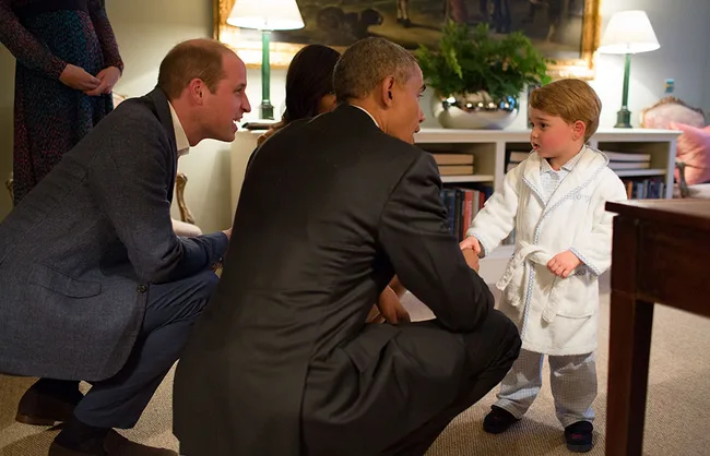 Three adults kneel to speak with a young boy in a bathrobe in a cozy, well-furnished living room.