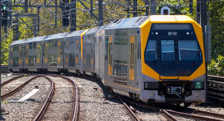 Yellow and grey train on tracks surrounded by overhead power lines and trees.