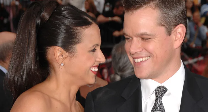 A couple smiling at each other at a formal event; the woman has a ponytail, and the man is wearing a suit with a patterned tie.