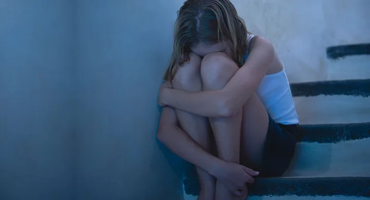 Young girl sitting on stairs, hugging knees, with head down, in a dimly lit environment.