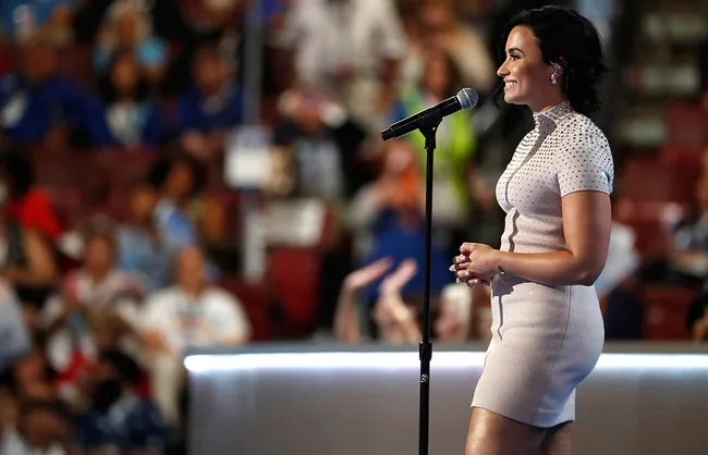 Singer in a light dress speaking at a microphone on stage at a crowded event.