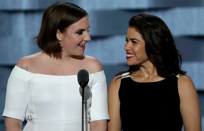 Two women smiling and standing close together, one in white off-shoulder dress, the other in a black dress, at a podium.