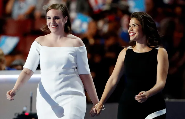 Two women on stage, one in a white dress and the other in a black dress, holding hands and smiling at an event.