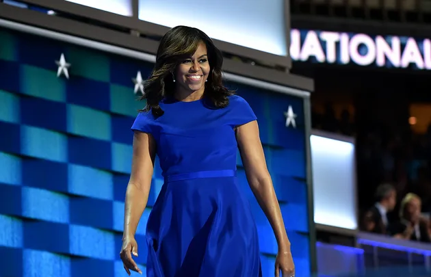 A woman in a blue dress smiles and walks on stage with a backdrop of blue, green, and stars.