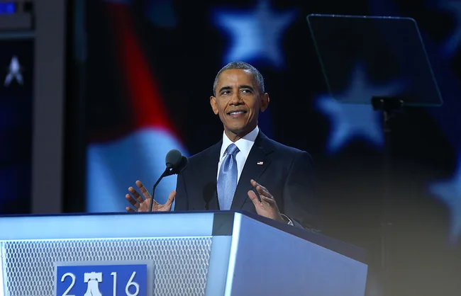 President speaking at a podium during the 2016 Democratic National Convention with a U.S. flag backdrop.