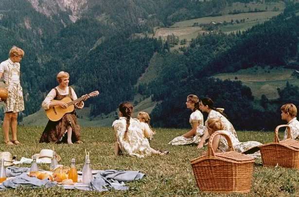 A woman playing guitar sings with children on a grassy hill, picnic baskets and food in the foreground, mountains in the background.