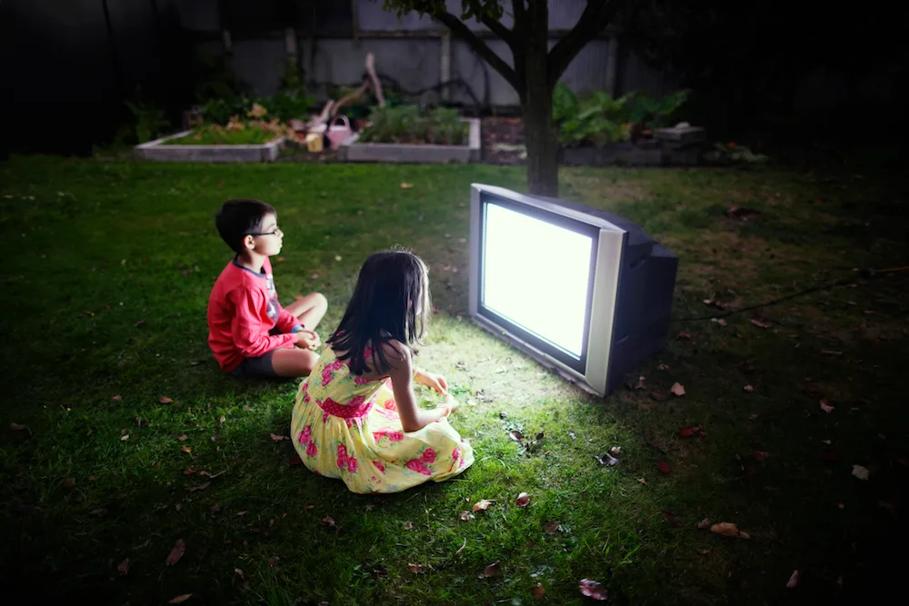 Two children sitting on grass at night, watching a bright TV screen outdoors.