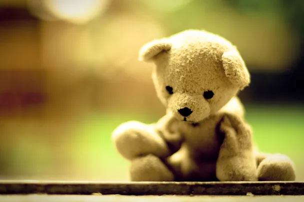 A small teddy bear sitting on a surface with a blurred green and yellow background.