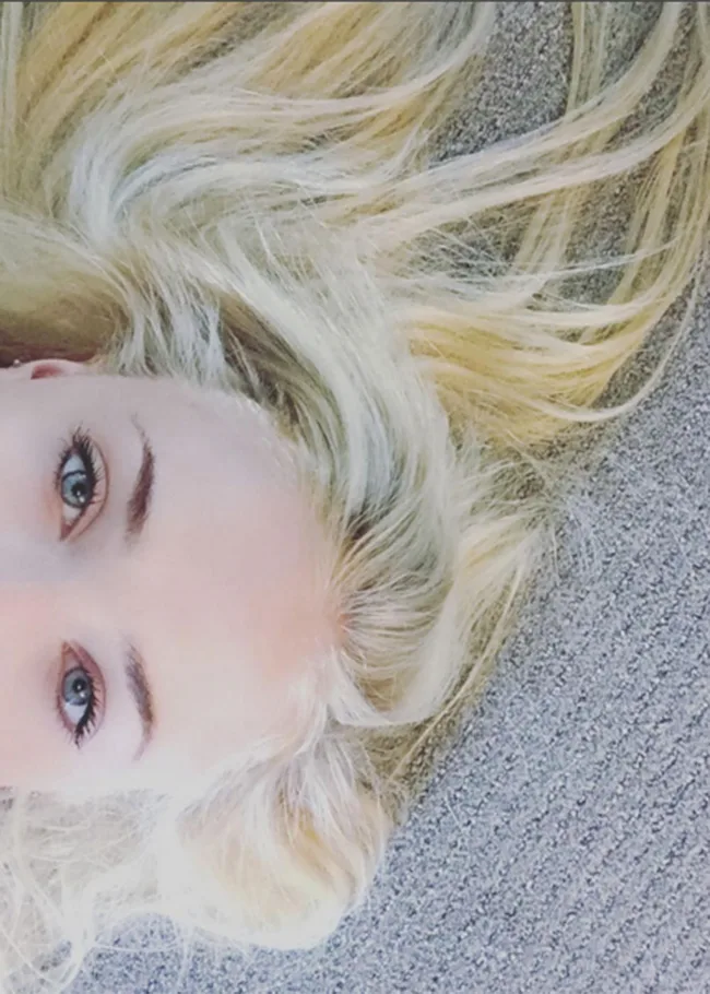Partial view of a person with long blonde hair and blue eyes lying on a gray surface.