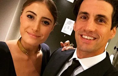 A woman in a black dress and a man in a suit smile for a selfie indoors.