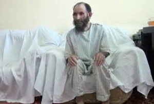 A bearded man in striped pajamas sits on a covered sofa in a sparsely furnished room.