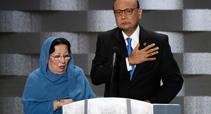 A man and woman stand at a podium; the man has his hand on his chest, the woman is wearing a blue headscarf.
