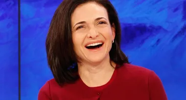 A person with short dark hair, wearing a maroon top, smiling against a blue background.