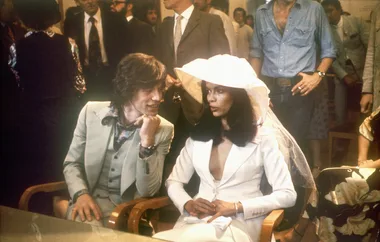A man in a light suit and woman in a white outfit with a hat sit together at an indoor event with people in the background.