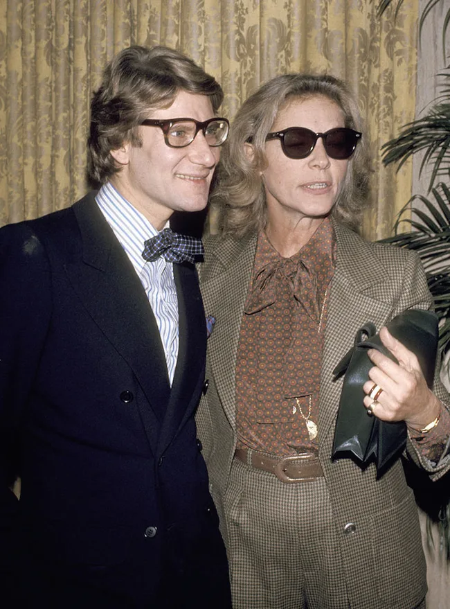 A man in a bow tie and glasses stands with a woman in sunglasses and a patterned suit, both smiling indoors.