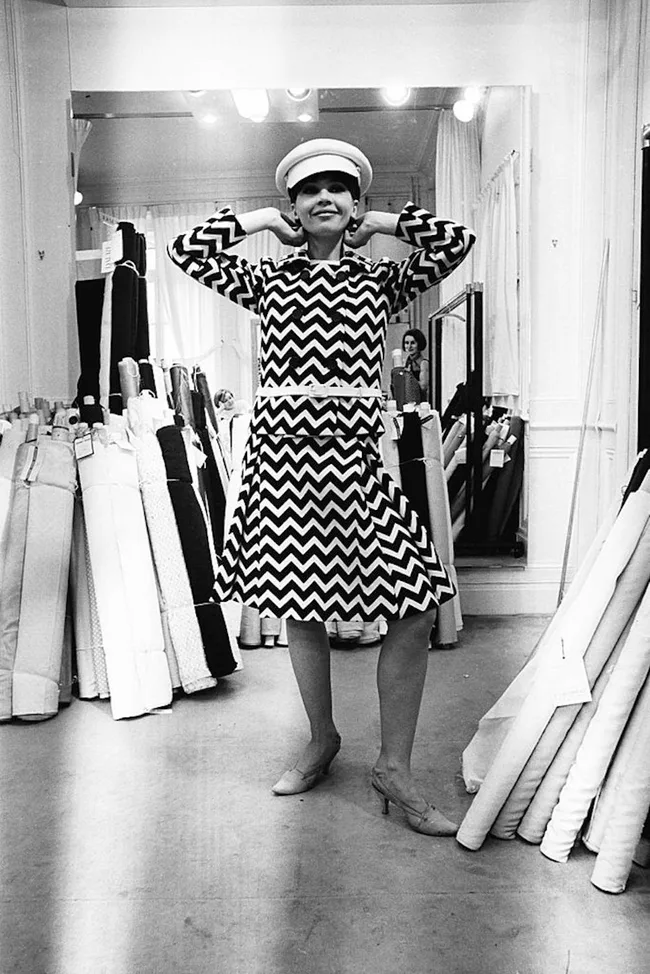 A woman in a zigzag-patterned dress and hat poses in a fabric store, surrounded by fabric rolls.