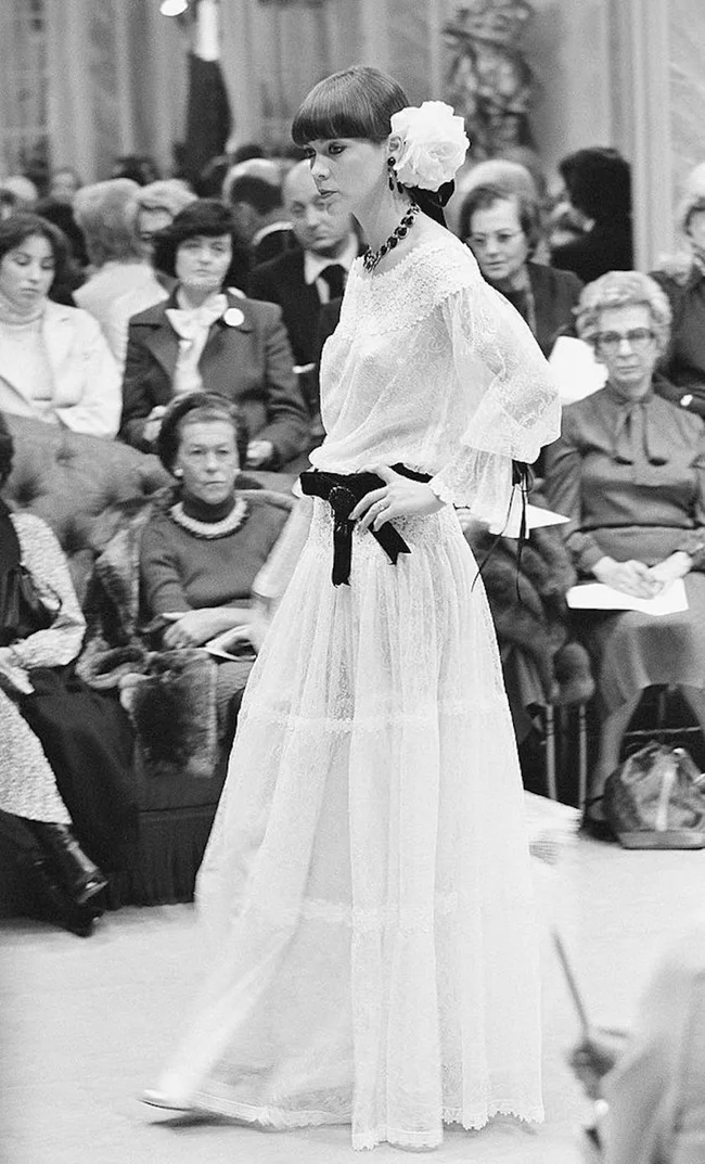 Model in a vintage white lace dress with floral headpiece walks at a fashion show, with seated audience in background.