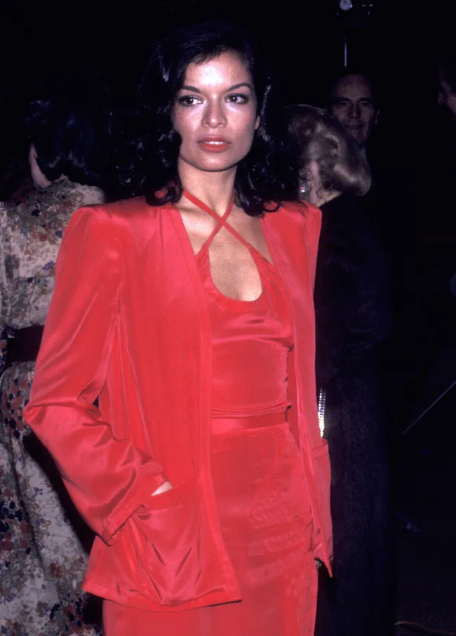 A woman in a vibrant red outfit with dark, wavy hair stands in a dimly lit event, looking off-camera.