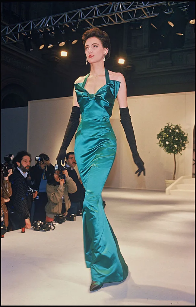 Model in teal satin gown with black gloves, walking a fashion runway surrounded by photographers.
