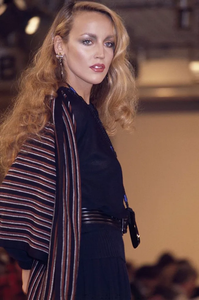 A person with long wavy hair wearing a black outfit and striped shawl on a runway.
