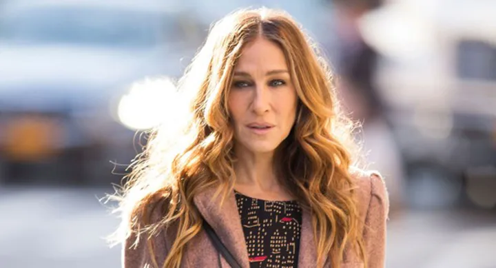 Woman with long wavy hair wearing a patterned top and coat, standing outdoors in soft lighting.