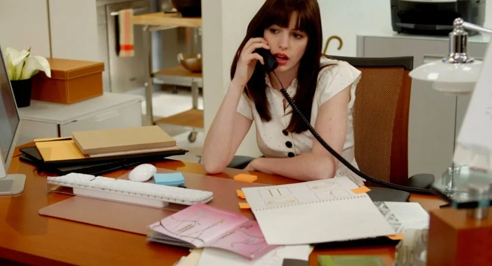 Young woman at a desk, talking on a phone, with papers and office supplies scattered around.