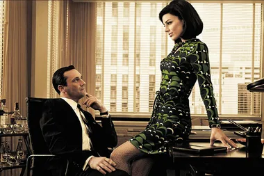 A man in a suit sits, looking at a woman in a patterned dress who is perched on a desk, with blinds in the background.