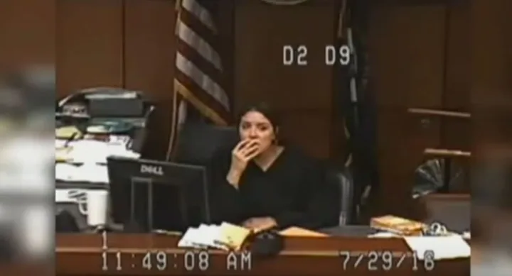 A judge in a courtroom, sitting at a desk with a computer and papers, appears to be listening attentively.