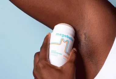 Person applying Megababe's Rosy Pits Daily Deodorant to their underarm against a light blue background.