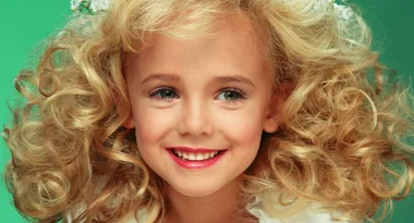 A young girl with curly blonde hair and a bright smile, dressed in white, against a green background.