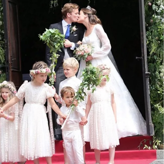 "Couple kissing at wedding, surrounded by children in formal attire, on a red carpeted entrance."