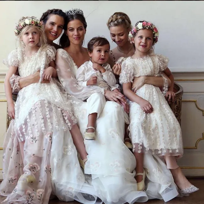 Women and children in matching white dresses and floral crowns sit together indoors, smiling and posing for a photo.