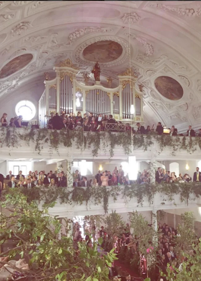 Elegant hall with ornate ceiling, lush greenery decor, a choir performing on upper balcony, and audience gathered below.