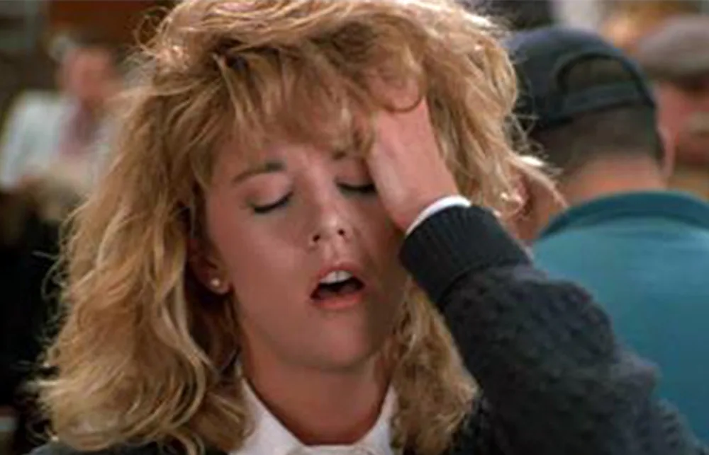 Woman with blonde hair and a dramatic expression holds her head in a crowded setting.