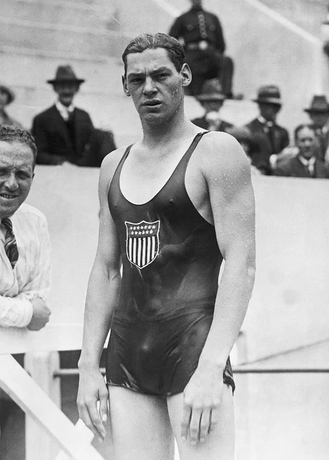 Male swimmer in vintage USA swimsuit stands poolside, onlookers and officials in the background. Black and white photo.