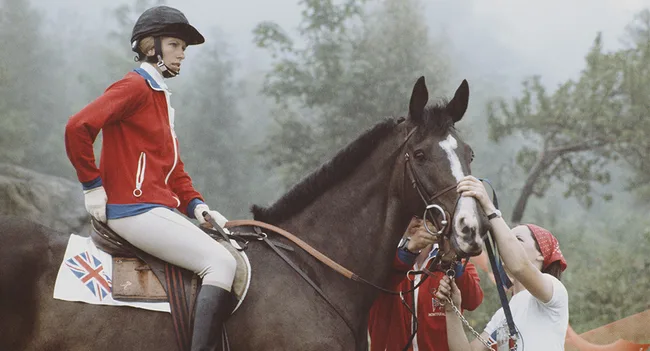 Rider in red jacket on horse with British flag saddle pad, assisted by a person with a red headscarf.