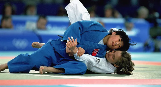 Judoka in blue gi holds opponent in white gi on mat during a match at the Olympics.