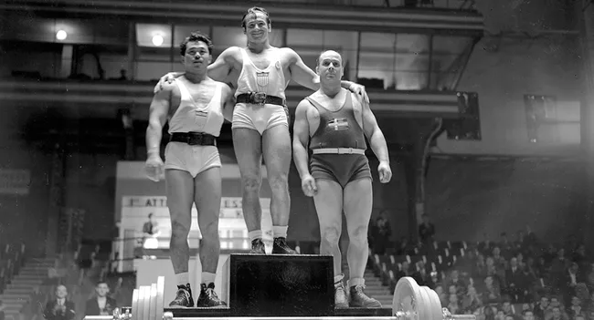 Three male weightlifters stand on a podium, celebrating a victory in an indoor arena.