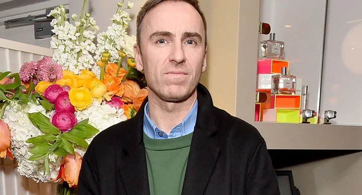 Man in a black coat stands in front of colorful flowers and perfume display.