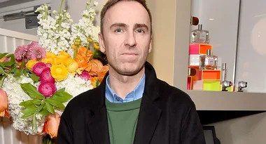 Man in a black coat stands in front of colorful flowers and perfume display.