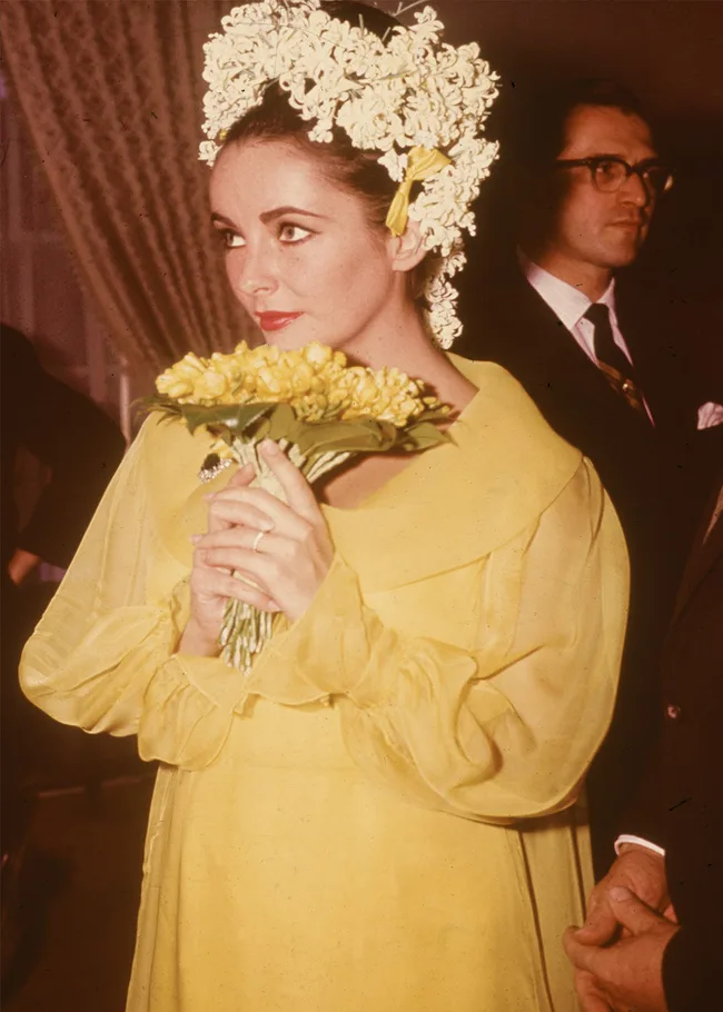 A woman in a yellow dress and floral headpiece holds a bouquet of yellow flowers, with a man in the background.