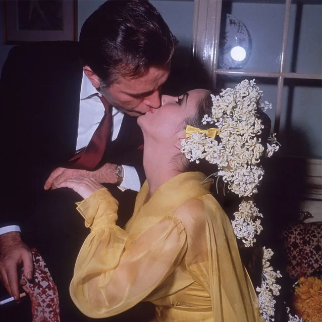 Couple shares a kiss; woman wears a yellow dress with floral headpiece, man in a suit with a red tie.